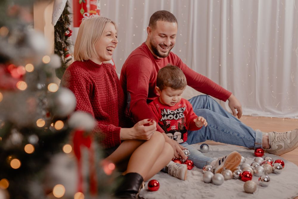 Happy family in red sweaters sitting on floor by Christmas tree with ornaments, laughing together during holiday celebration.