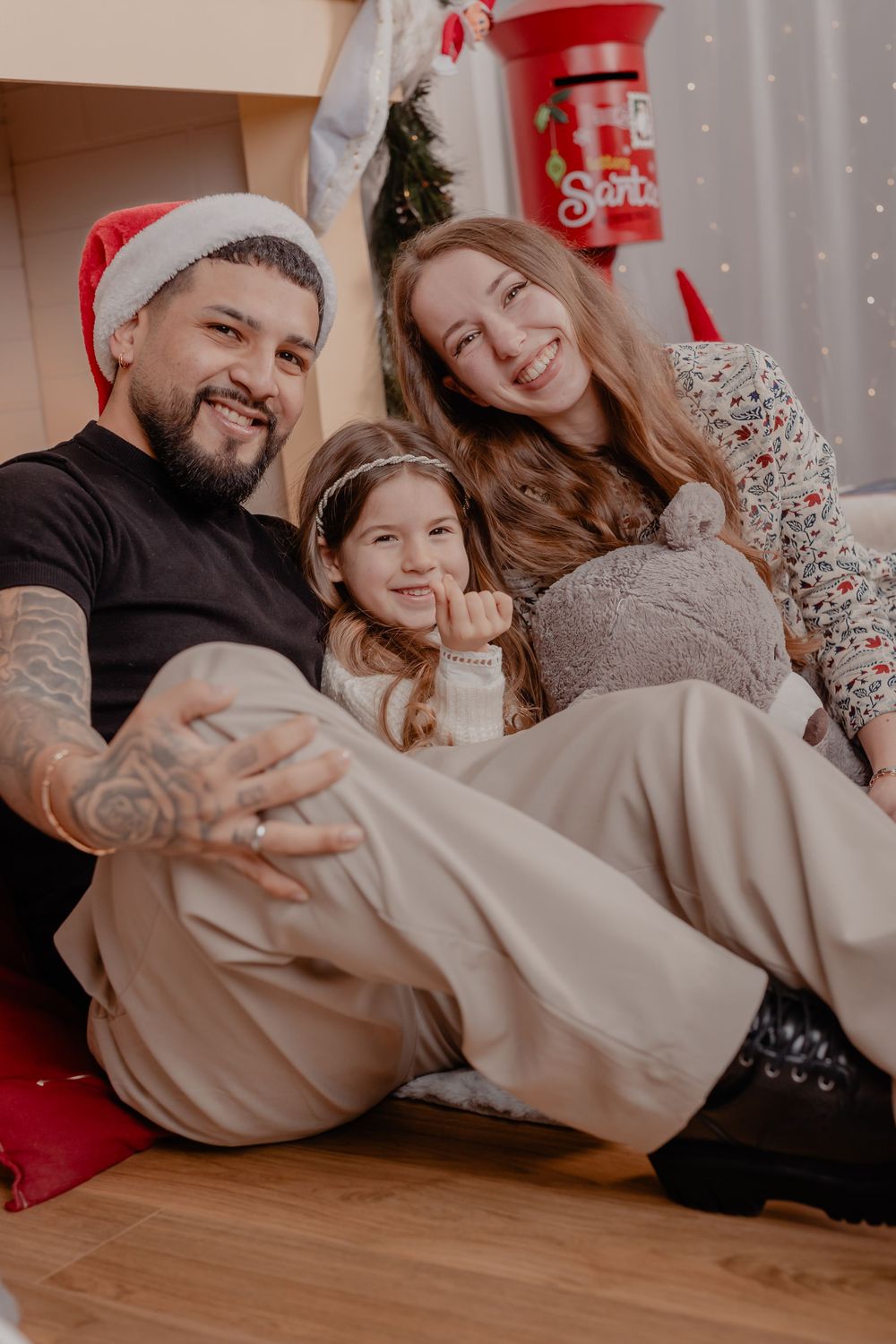 Happy family with tattooed dad in Santa hat, mom, and daughter posing together in front of Christmas decorations.