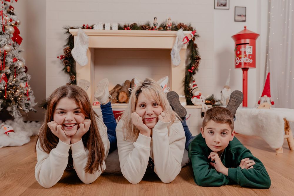 Three children pose cheerfully on the floor in front of a decorated fireplace and Christmas tree in a festive holiday setting.