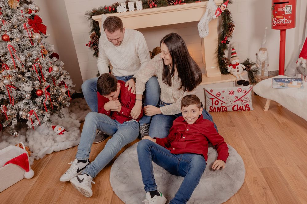 Family of four sitting by decorated Christmas tree and fireplace, wearing red and white holiday outfits.