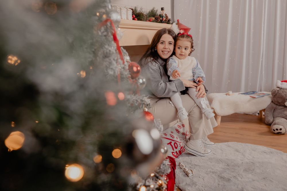 Woman and toddler girl sitting together by a decorated Christmas tree and fireplace in a cozy holiday setting.