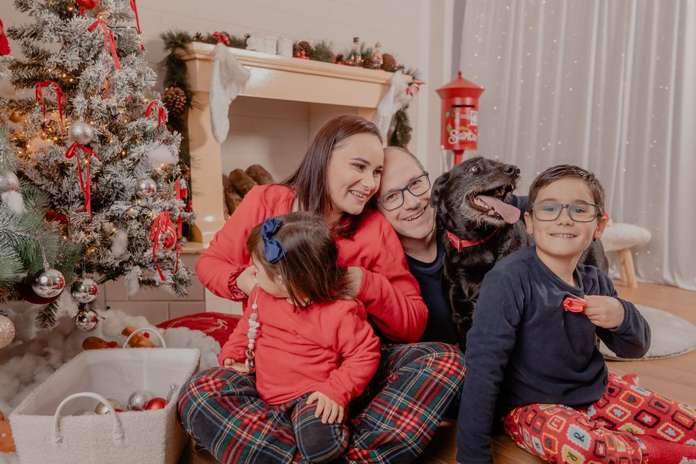 Family in red plaid pajamas smiling with black dog near decorated Christmas tree and fireplace.