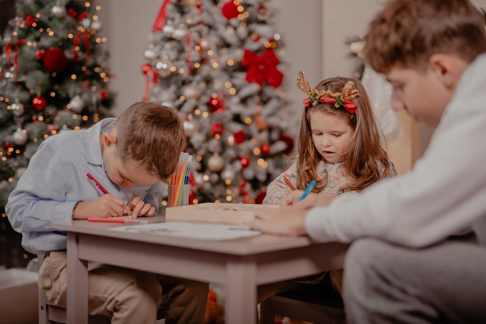 Three children writing letters to Santa Claus at a small table near a decorated Christmas tree.