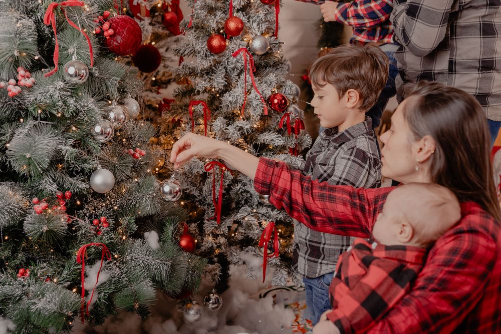 Children in plaid shirts decorating a Christmas tree with red ornaments while holding a baby during holiday celebrations.