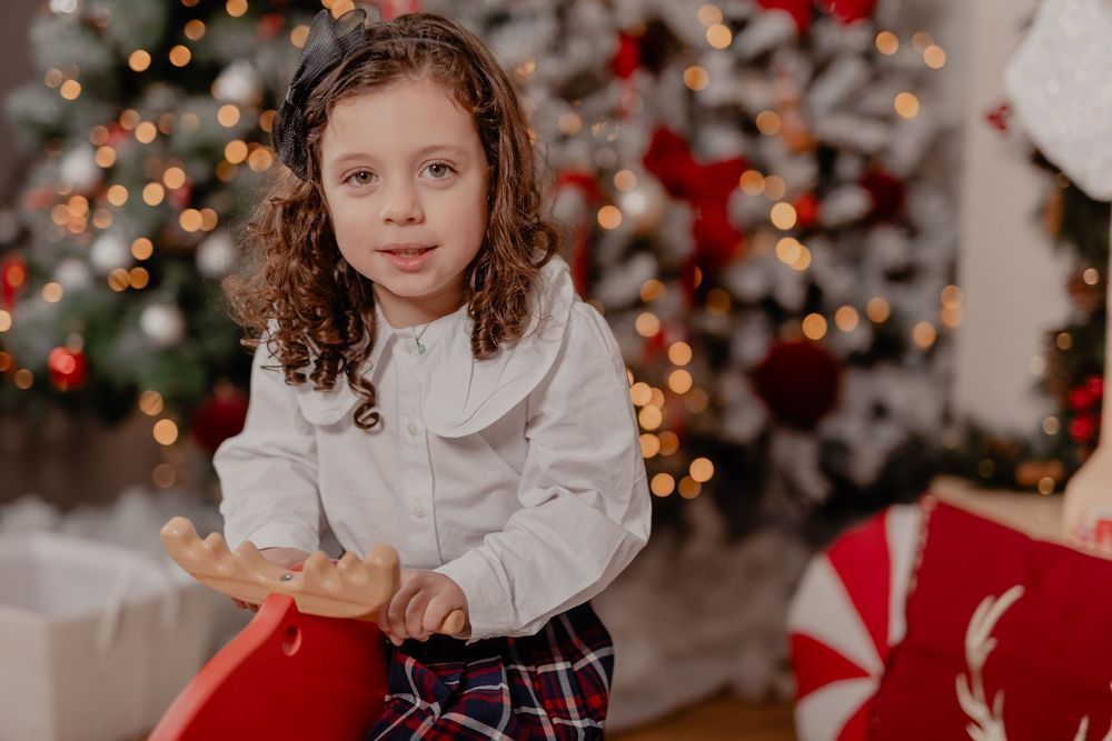 Young girl in white blouse and plaid skirt poses near decorated Christmas tree with festive red and white decor.