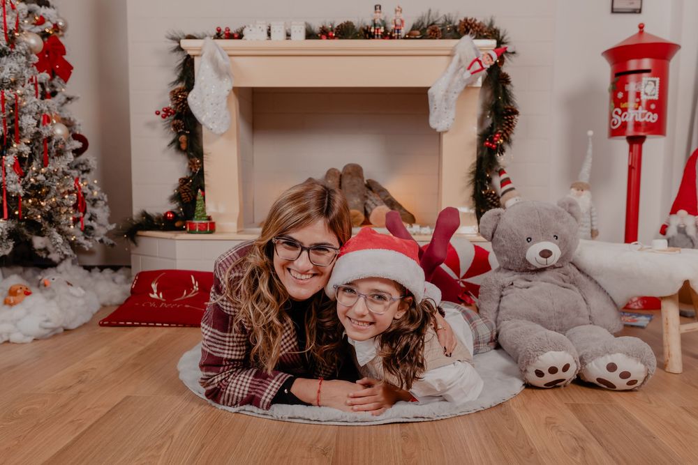 Mother and daughter smiling together by a decorated Christmas fireplace with a teddy bear and holiday tree.
