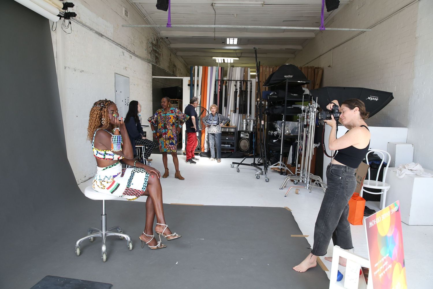 Photographer taking photos of a woman sitting on a stool in a professional photography studio with lighting equipment.