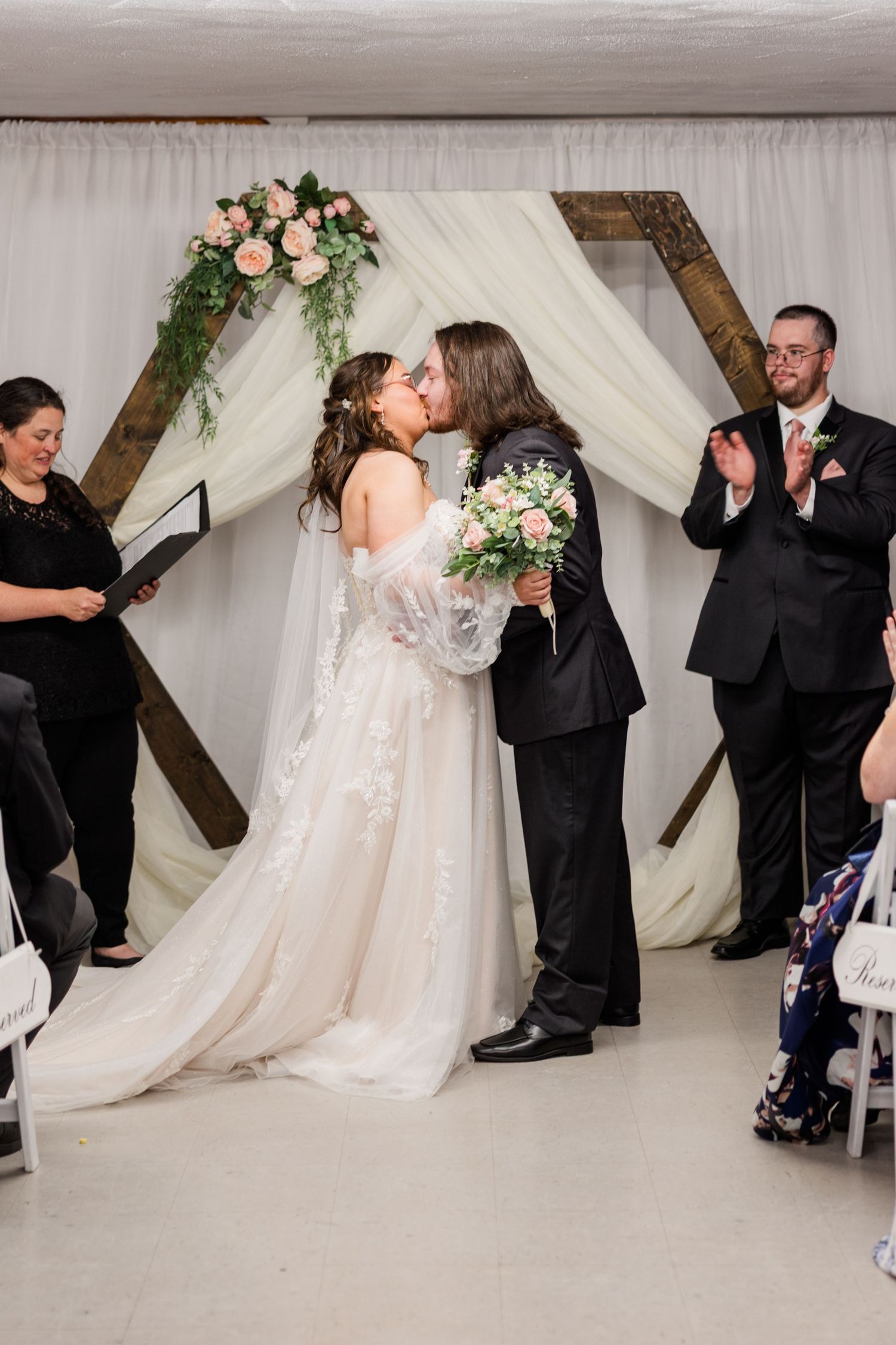 Bride and groom share first kiss at altar with wooden hexagon arch, pink flowers, and white draping at indoor wedding ceremony.