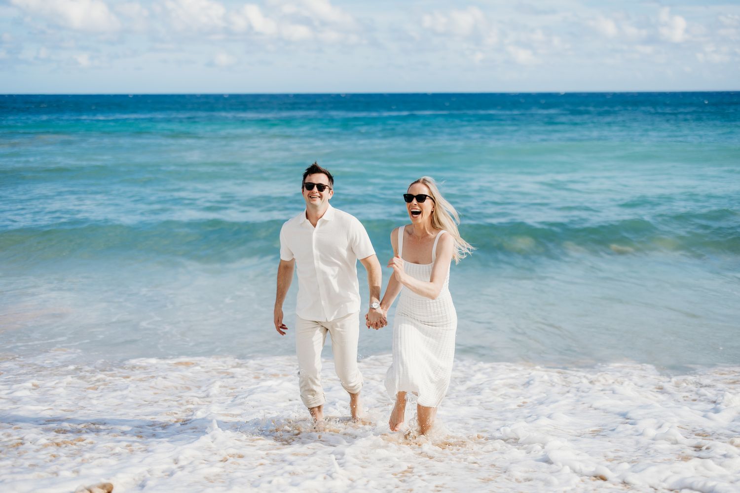 Happy couple in white outfits holding hands running on a tropical beach.