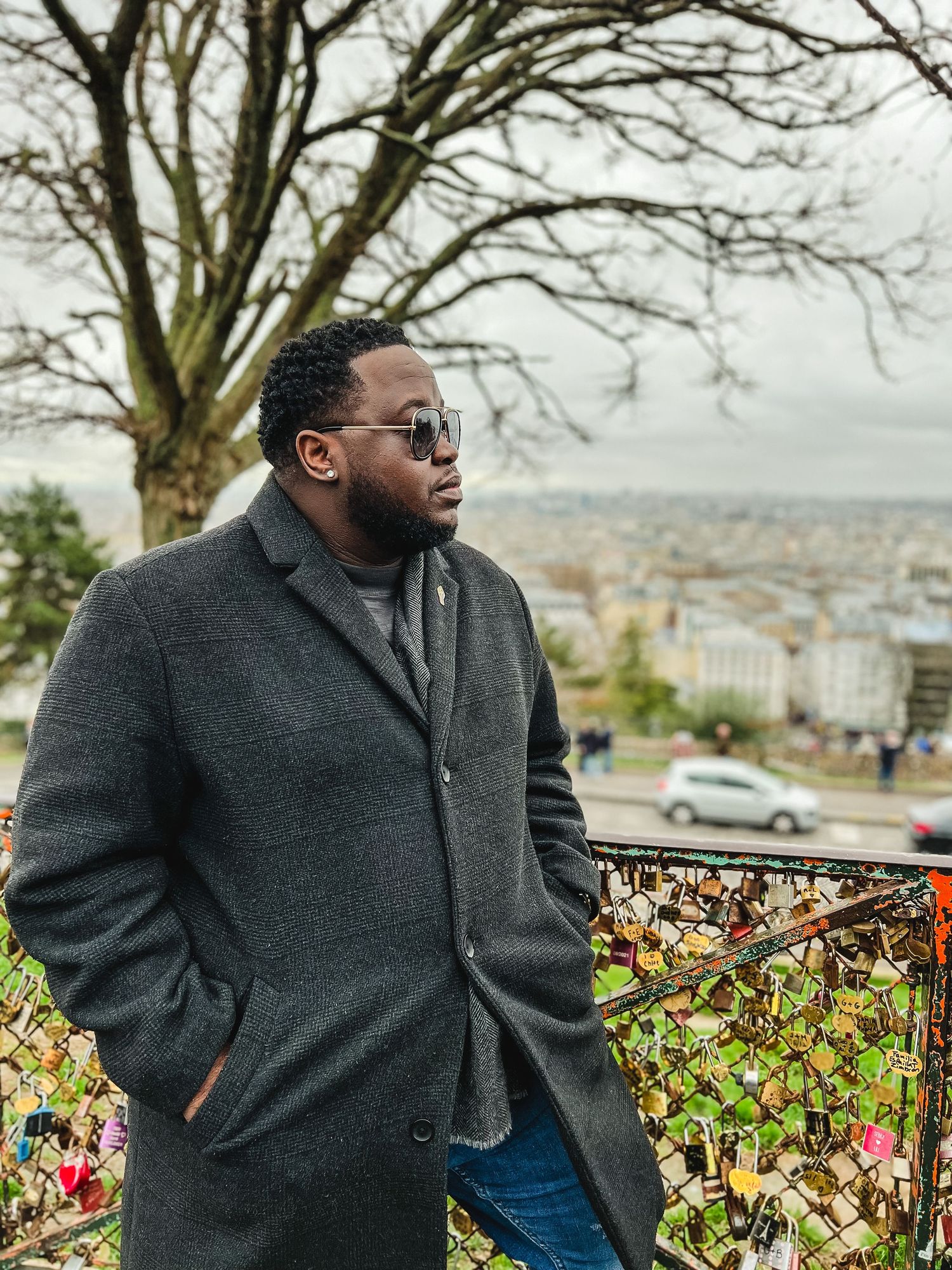Man in sunglasses and a gray wool overcoat stands beside a fence covered in love locks, overlooking a hazy city skyline and bare tree.