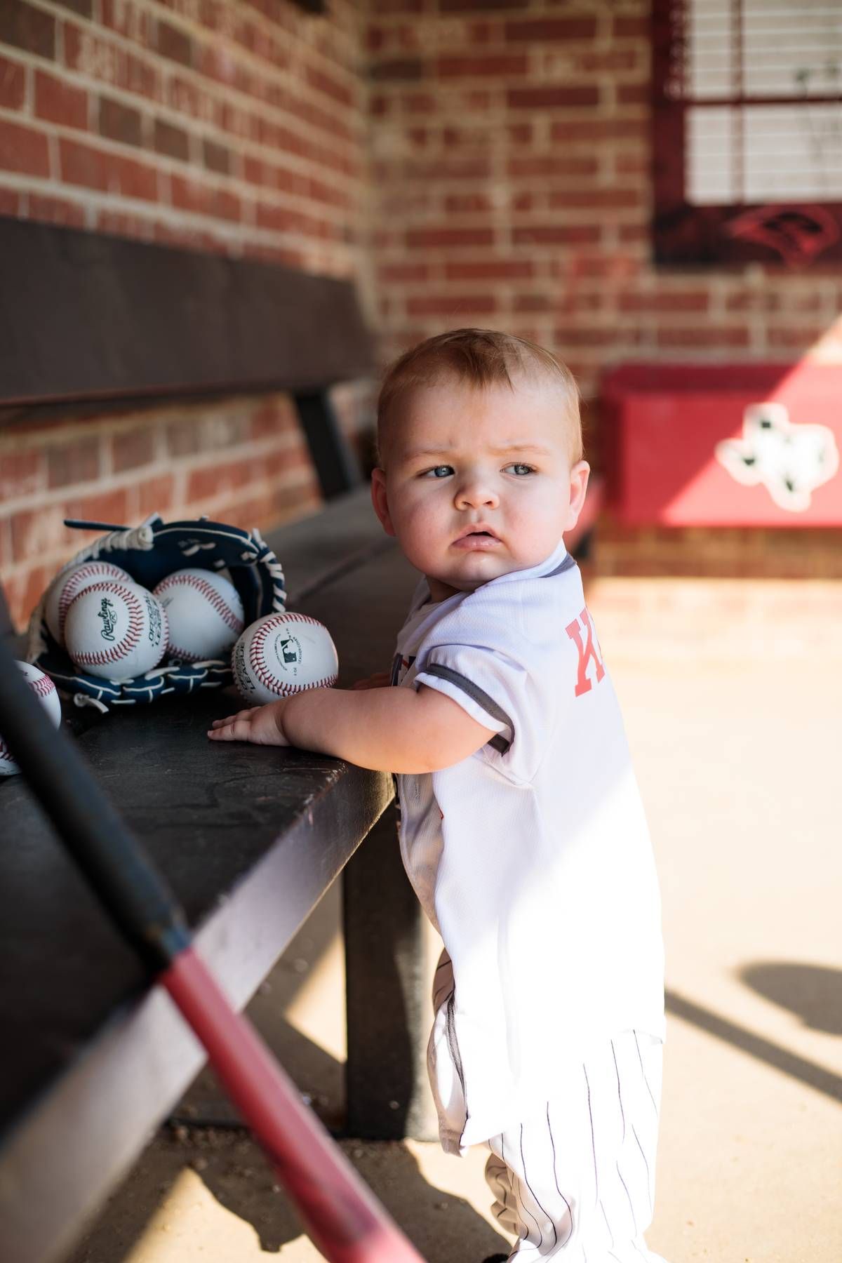 Baby in a white pinstripe baseball uniform stands in a dugout beside a bench with baseballs, a glove and a red bat.