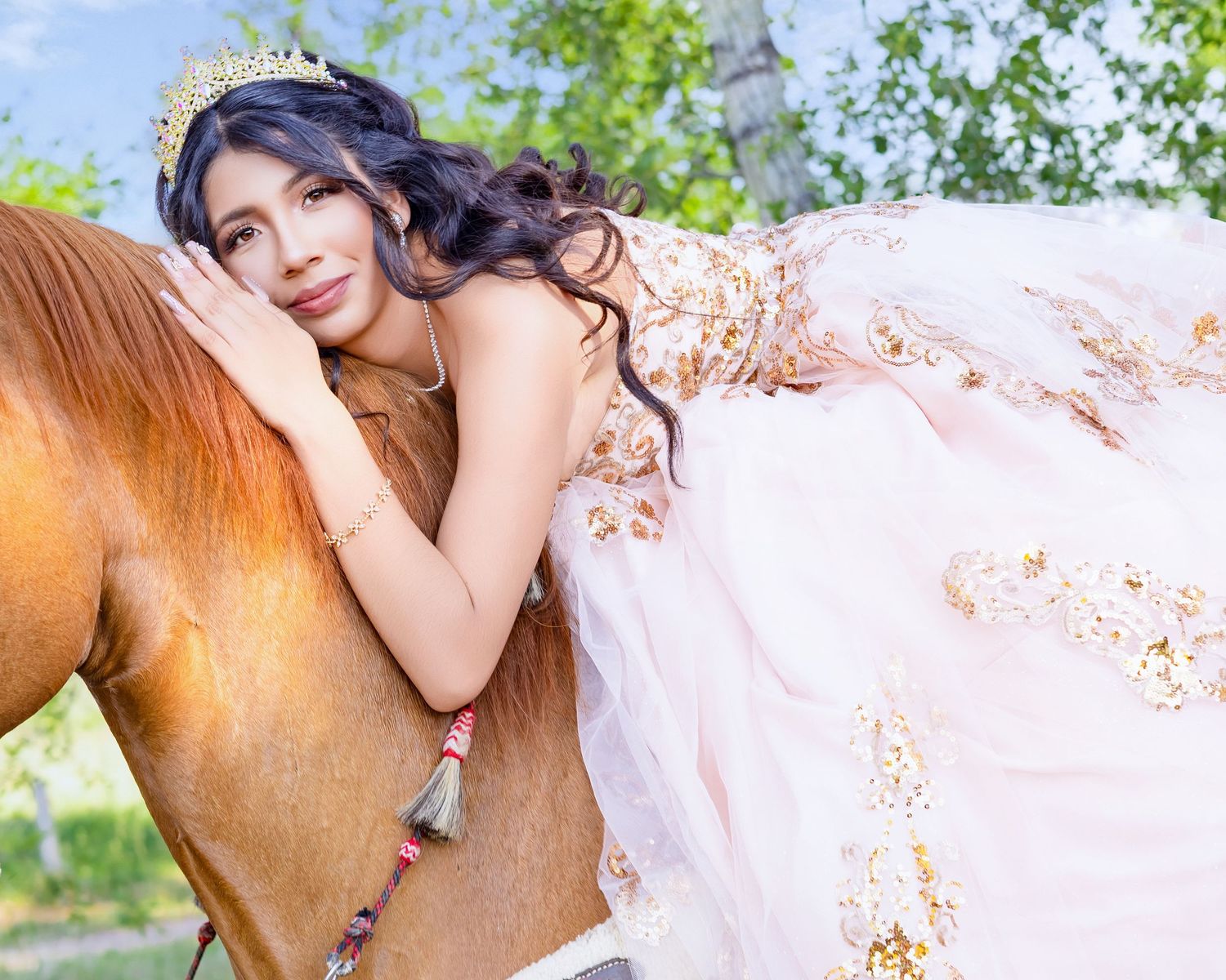 Young woman in pink floral quinceañera gown and crown leans gracefully against a chestnut horse outdoors.