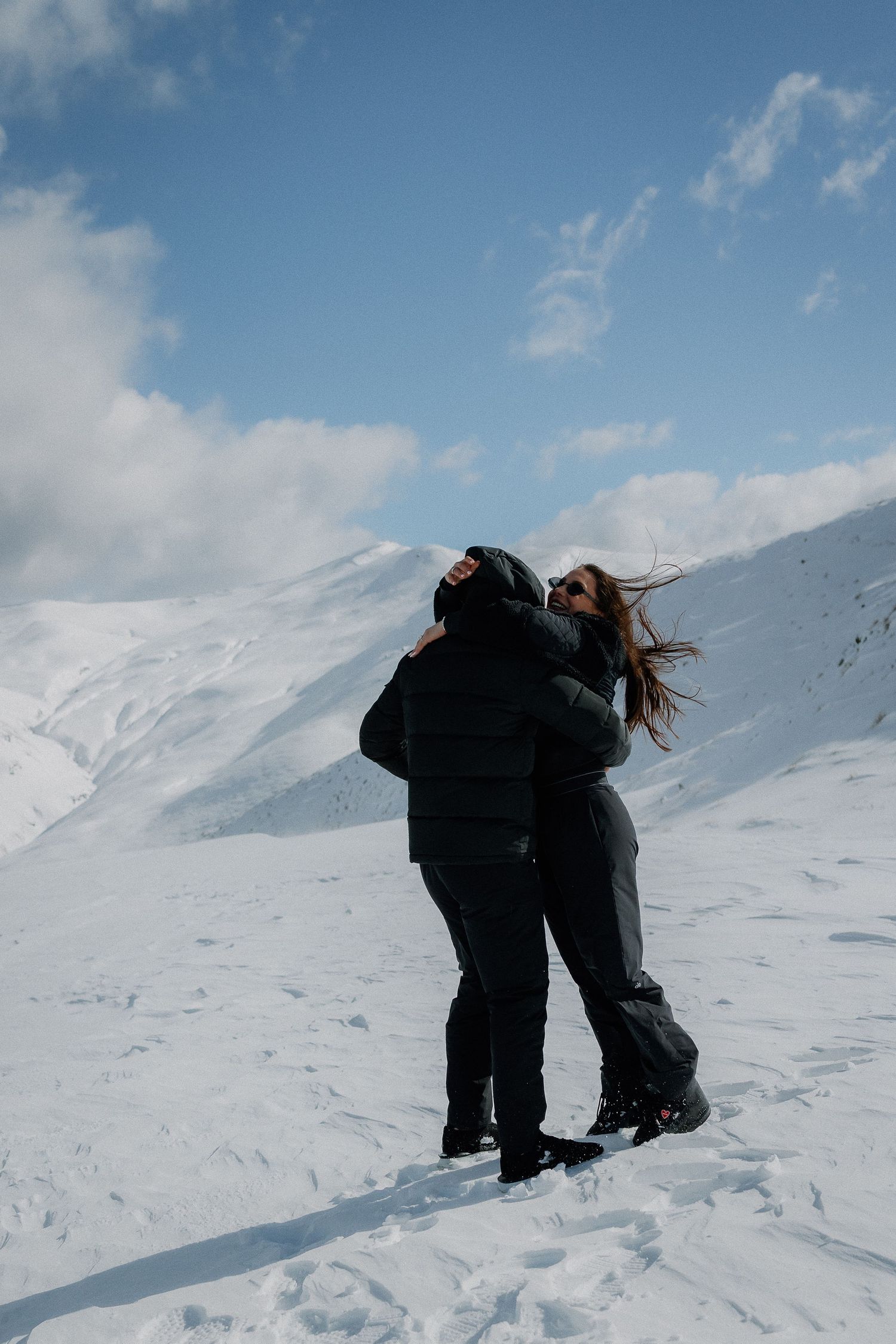 Couple embracing on a snowy mountain slope under a blue sky, woman's hair blowing in the wind.