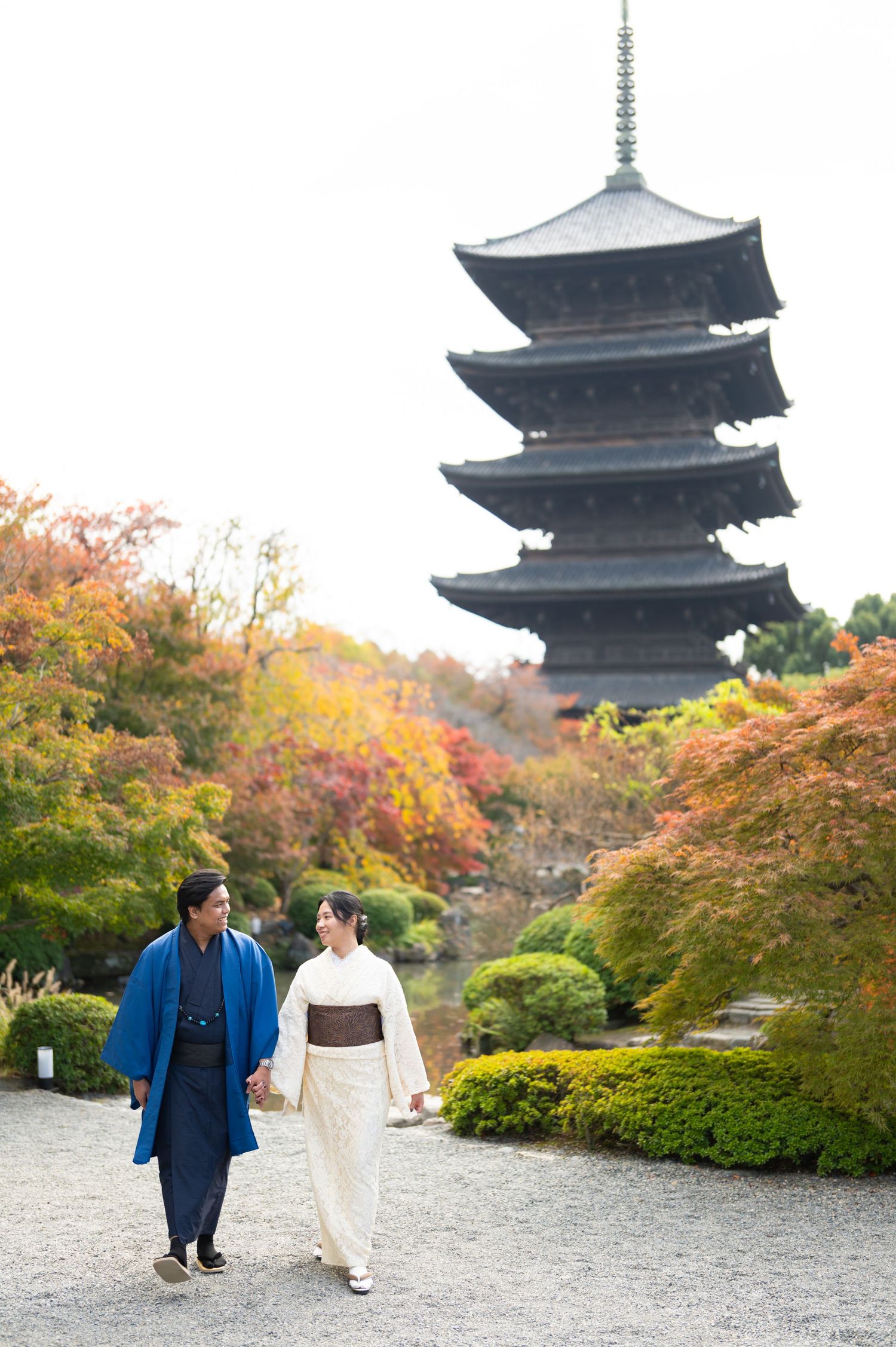 Couple in traditional Japanese kimono walking hand in hand through a garden with colorful autumn foliage and a historic five-story pagoda.