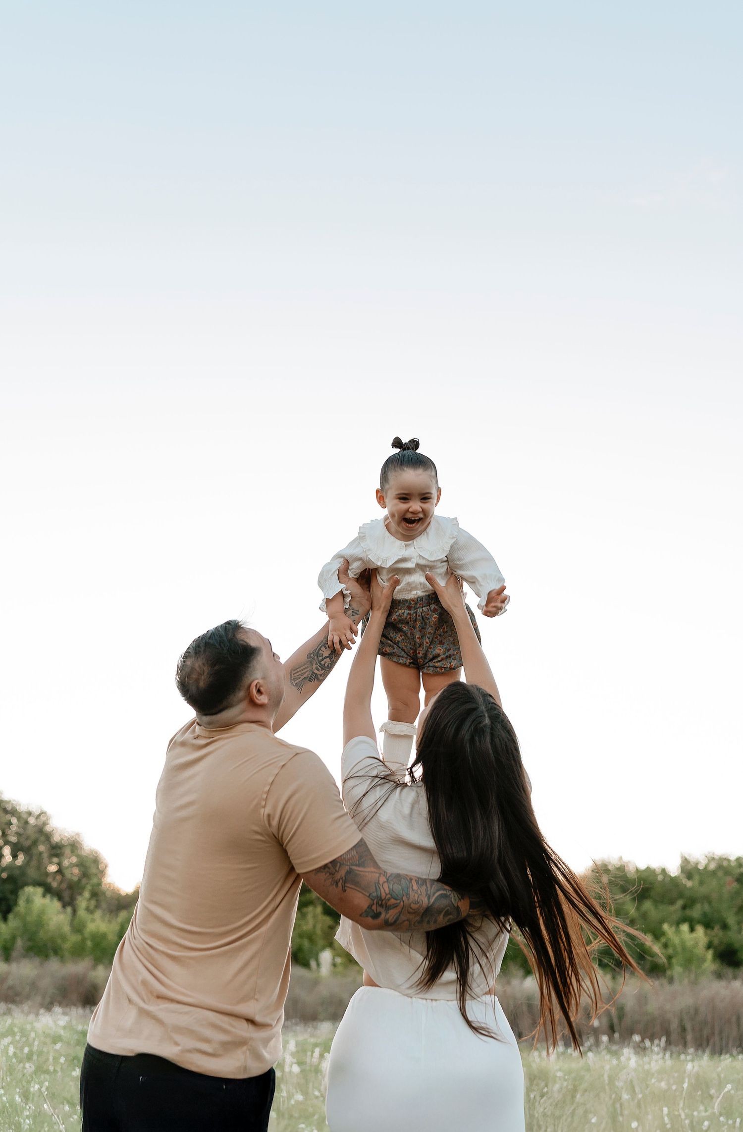 Parents joyfully lifting their laughing baby girl in the air during an outdoor family photo session in a green meadow.