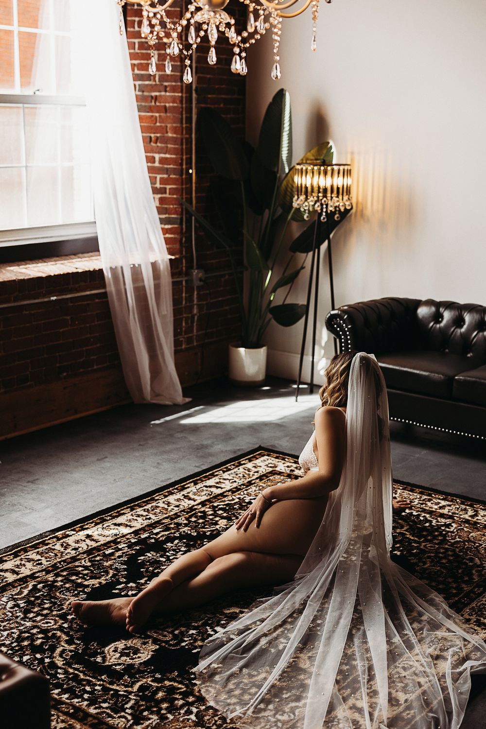Bride in a wedding veil seated on an ornate rug in a luxurious loft with brick walls and crystal chandelier.