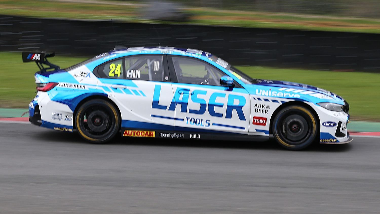 BTCC racer number 24 drives a white Laser Tools branded car at speed on a motorsport circuit.
