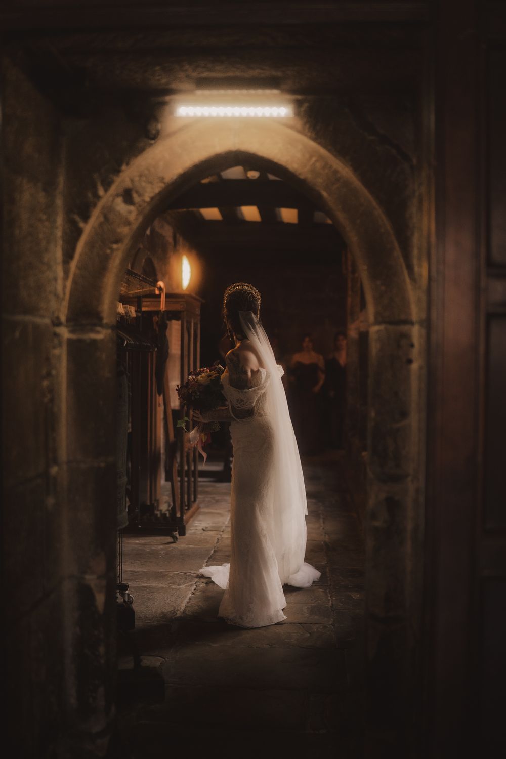 Bride in white gown standing in a stone archway of a dimly lit historic church, holding a bouquet.