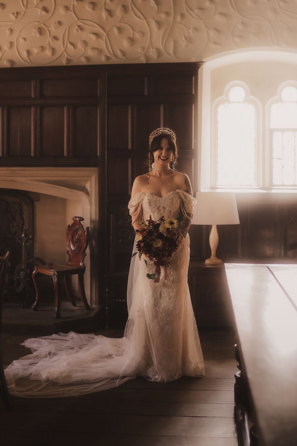 Bride in off-shoulder lace gown with tiara holds bouquet in elegant dark wood room with arched windows.