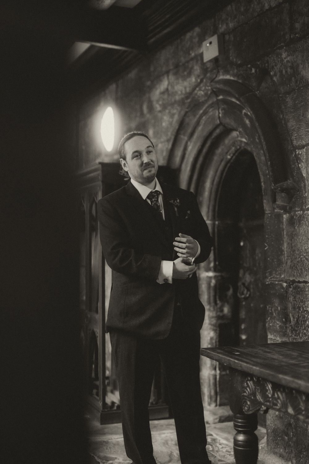 Black and white photo of a groom in a suit adjusting his cufflinks inside a historic stone church.