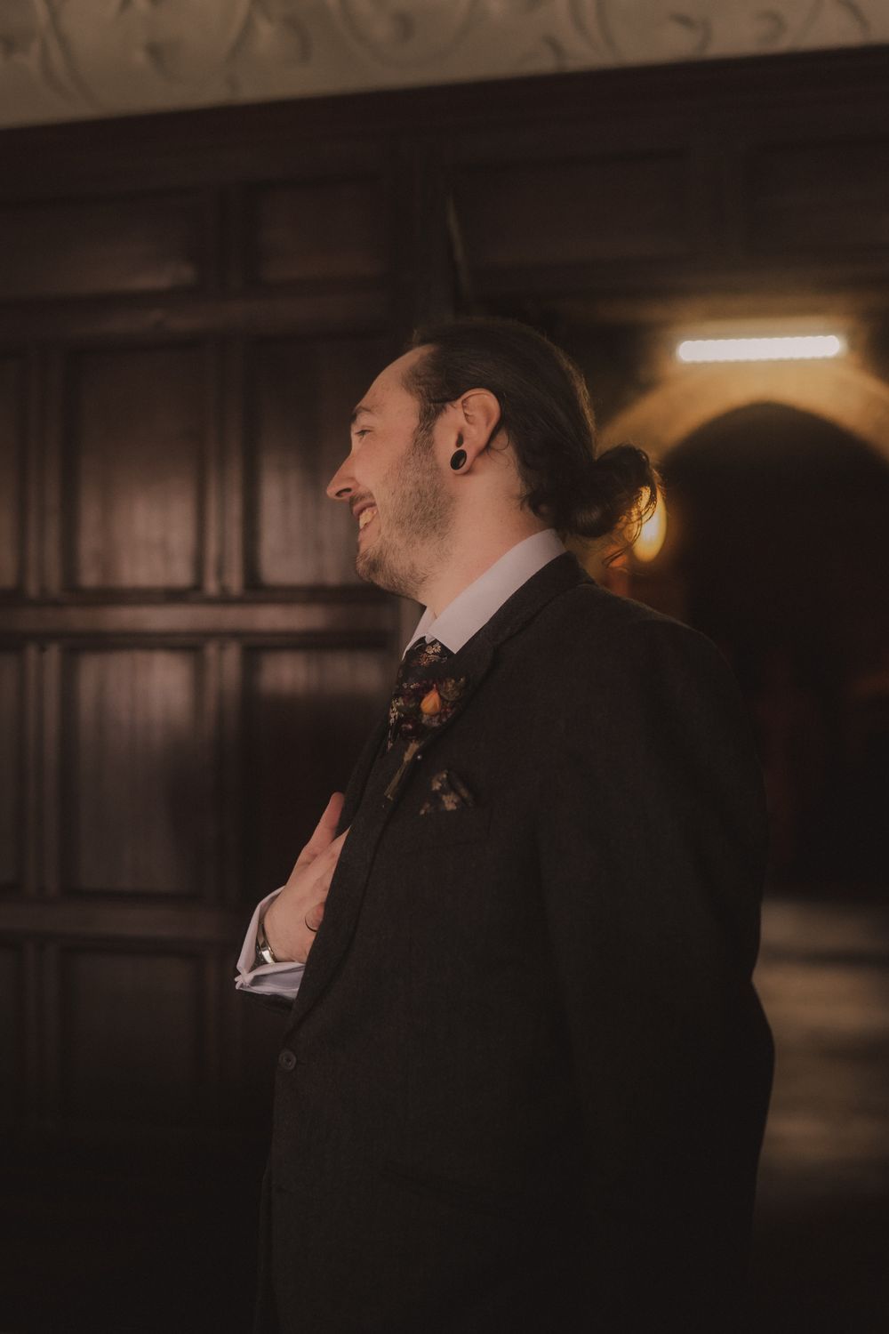 A stylish groom in a dark suit with floral bow tie and pocket square, smiling in a dimly lit elegant venue.