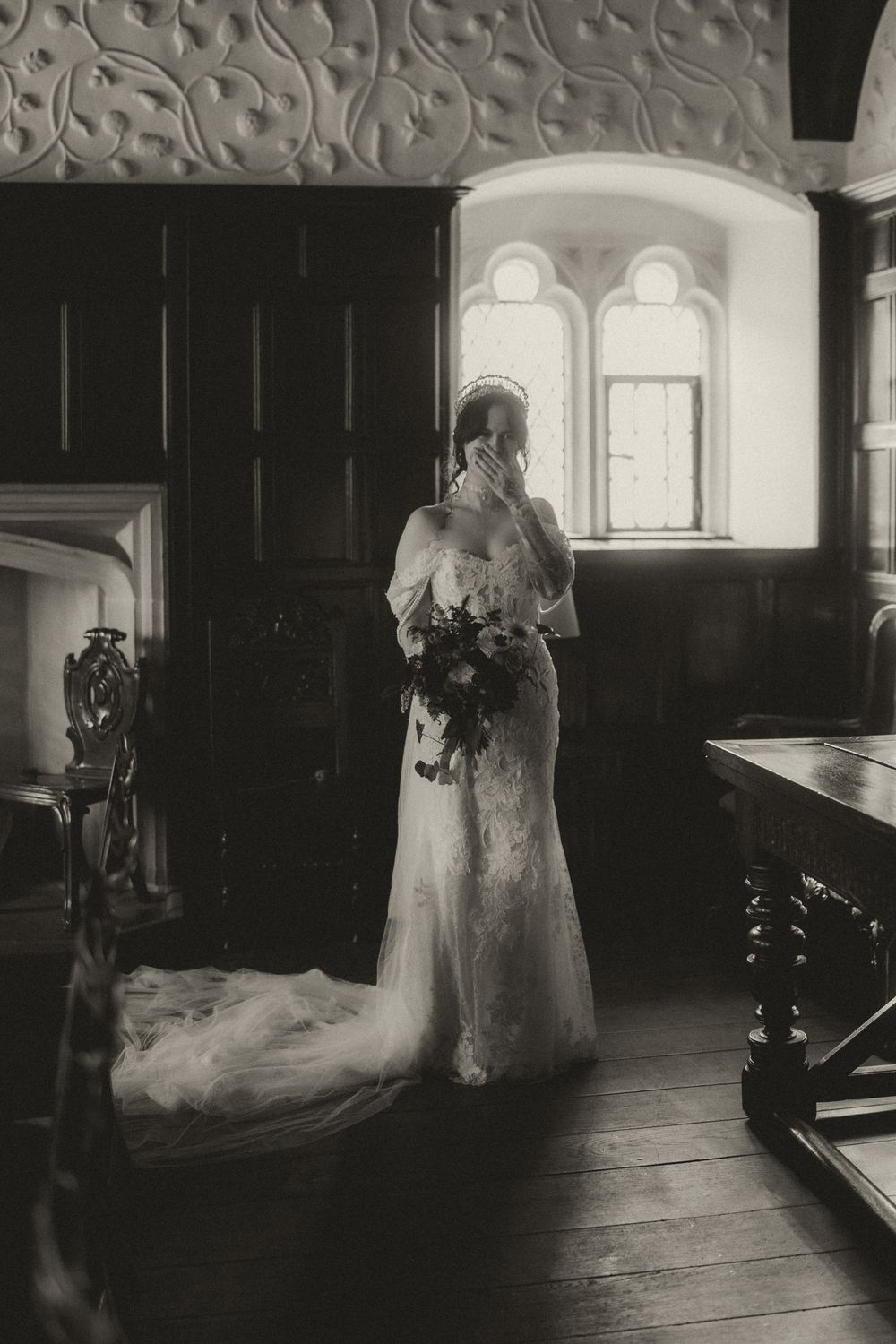 Black and white photo of a bride in a lace gown holding a bouquet, standing by arched windows in a historic manor.