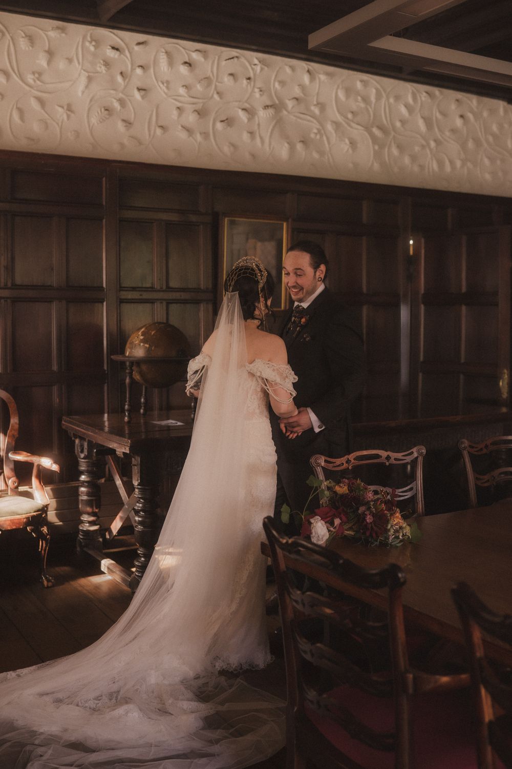 Bride and groom share a romantic moment in an elegant wood-paneled room, her long veil trailing gracefully on the floor.