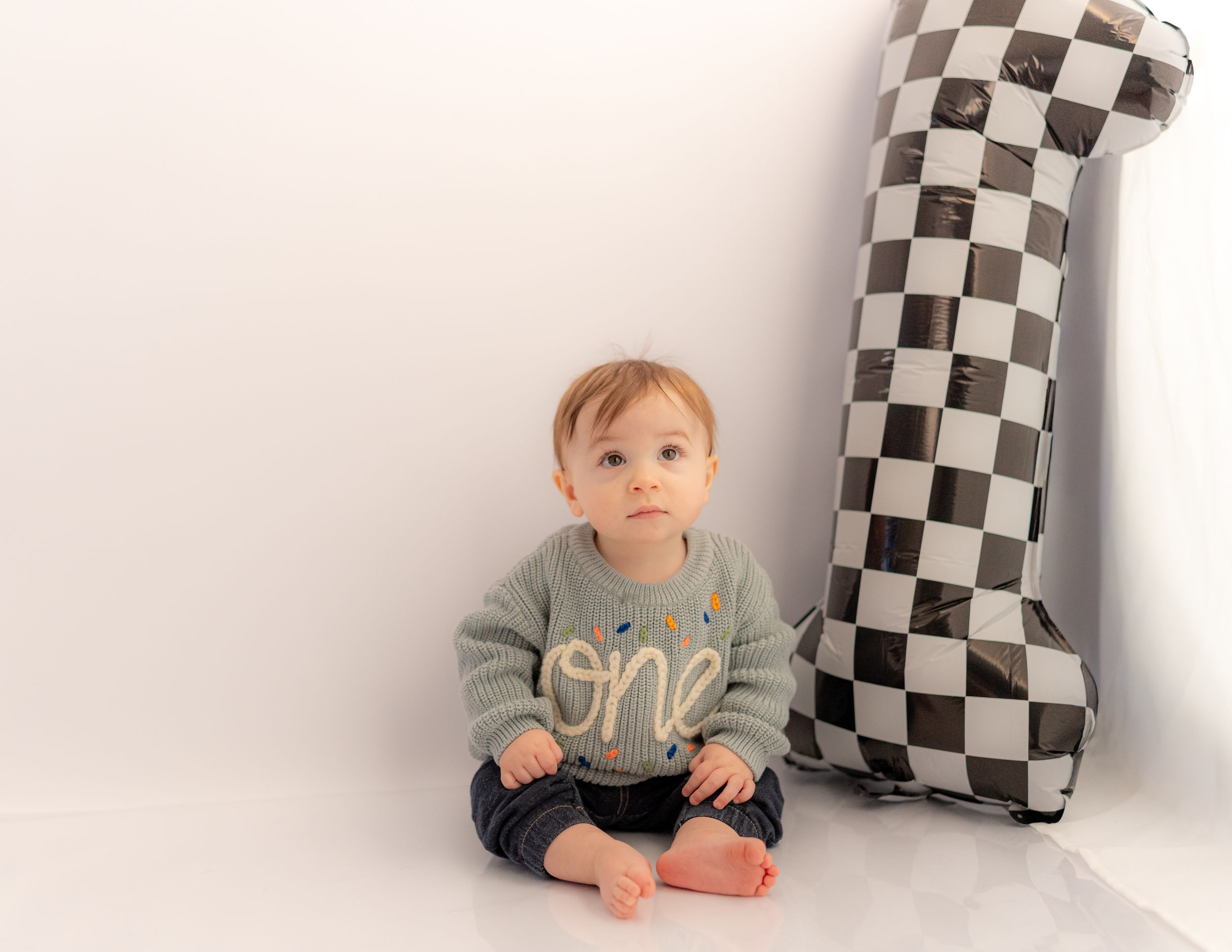 Baby in a gray ‘one’ sweater sits beside a checkered number 1 balloon celebrating their first birthday.
