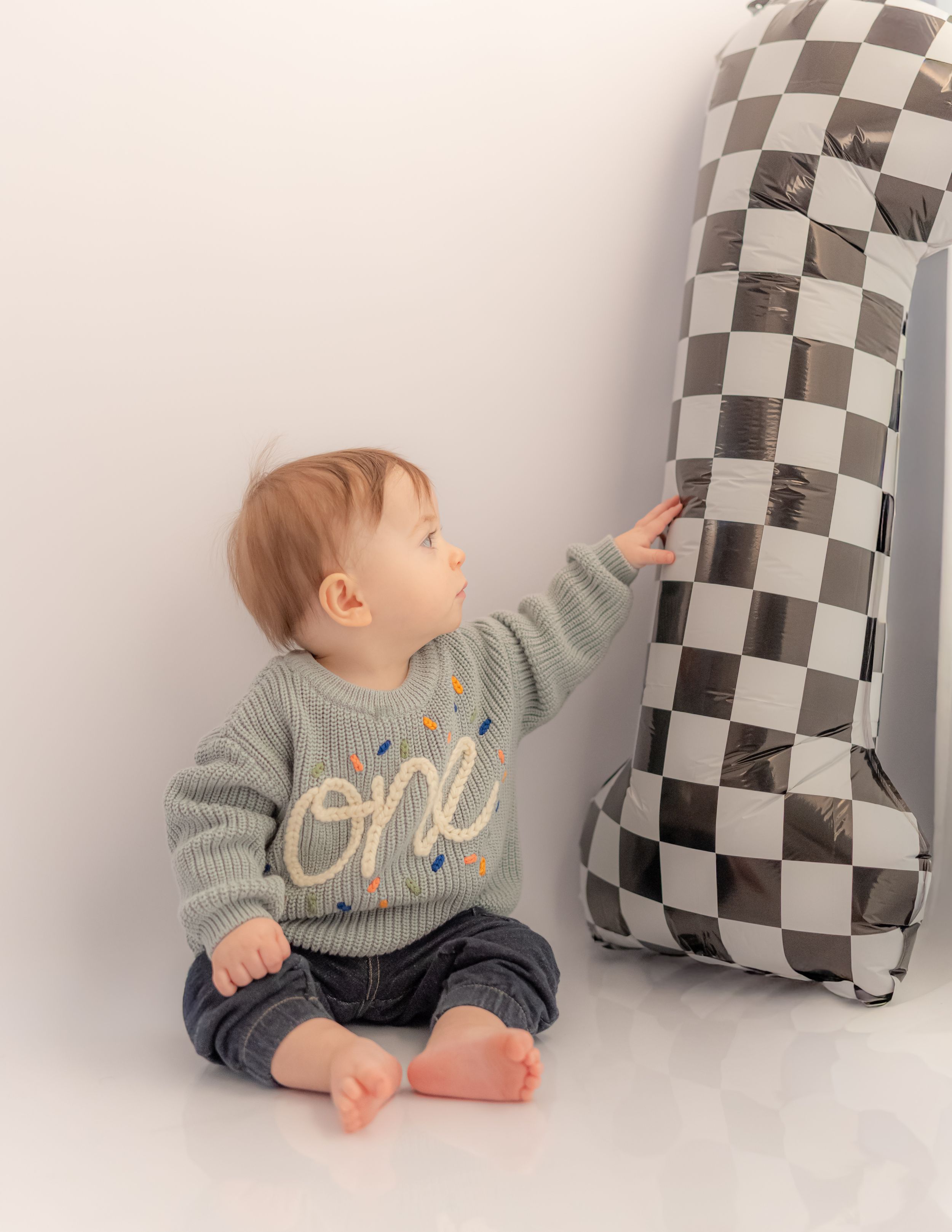 Baby in 'one' sweater touches checkered number 1 balloon, celebrating their first birthday.