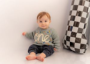 Adorable baby wearing a gray 'one' sweater sits smiling on white backdrop next to checkered pillow.