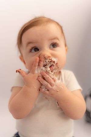 A messy baby with cake-covered hands and face, enjoying their first birthday smash cake on a white background.