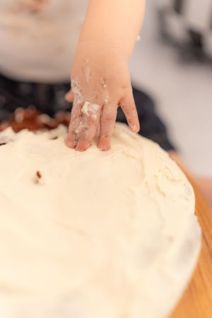 Child's hand pressing into white frosting on a cake, with cream-covered fingers during cake decorating.