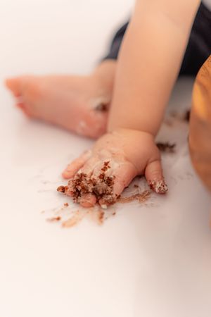 Baby hand and feet covered in chocolate cake crumbs during first birthday cake smash on white background.