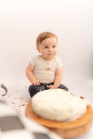 A messy baby in a white onesie sits next to a white frosted cake during a first birthday cake smash photo session.