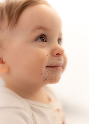 Adorable baby with chocolate smeared around mouth looking up, wearing white outfit against light background.