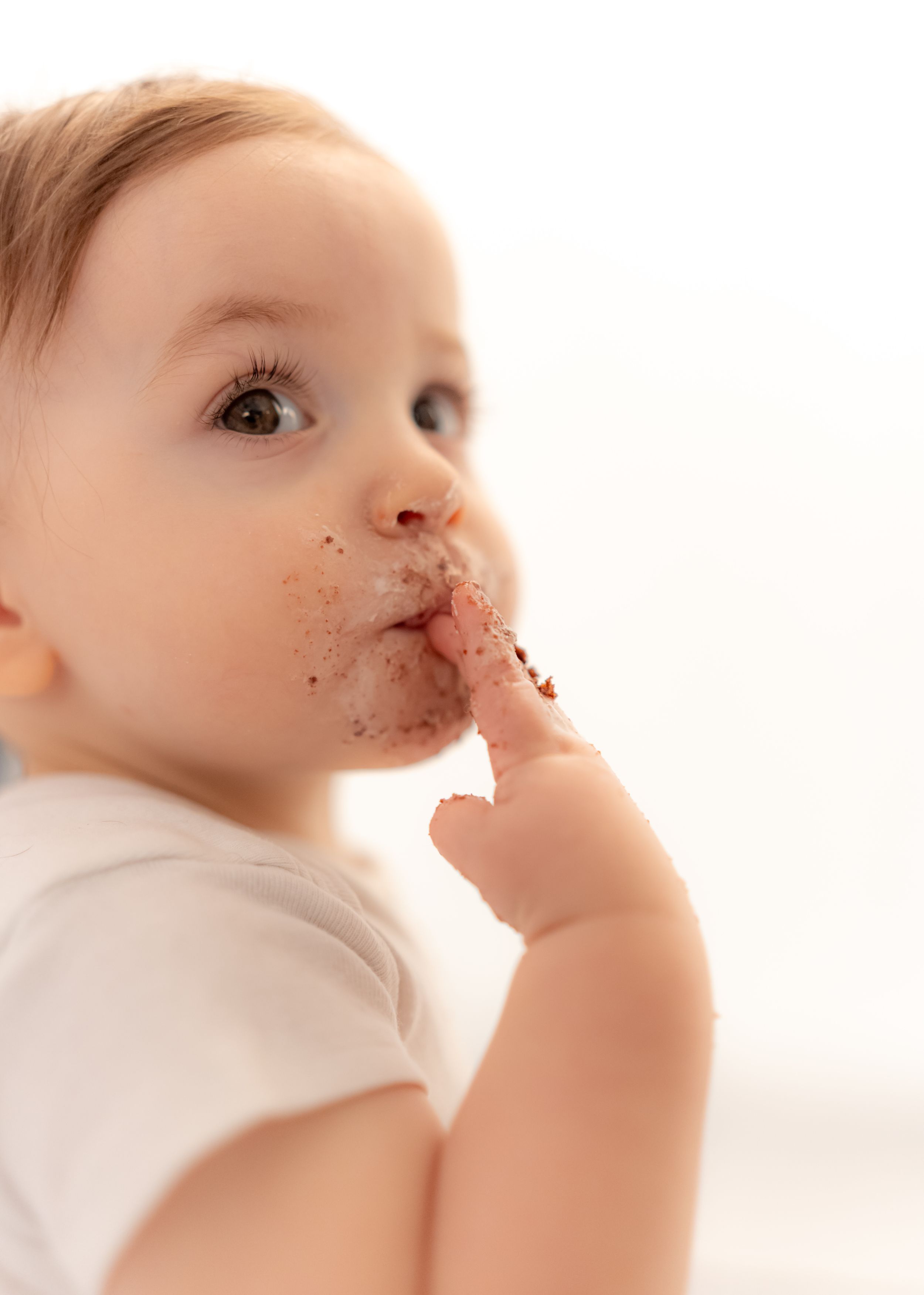 Cute baby with chocolate-covered face licking finger, wearing white onesie against light background.