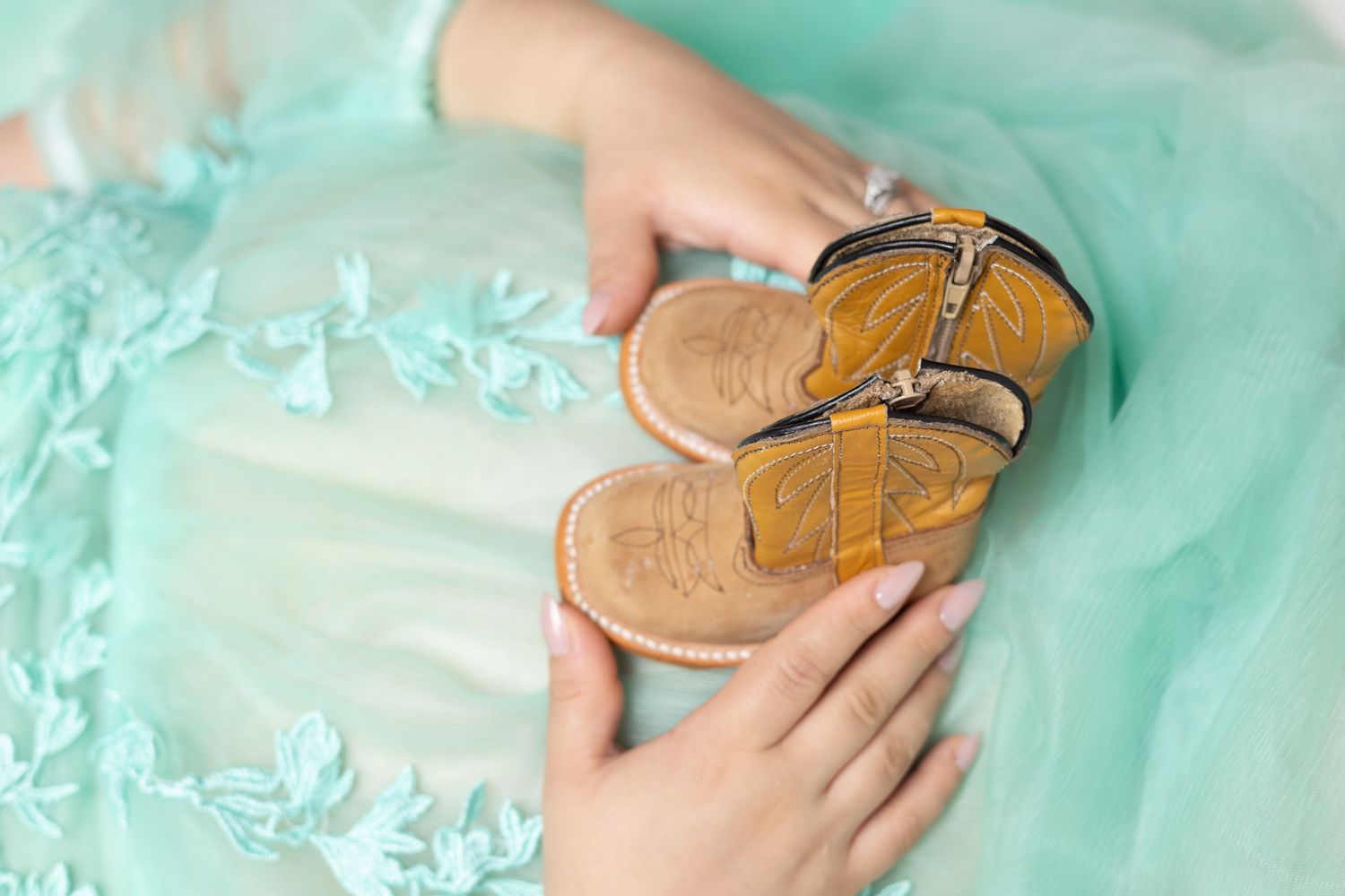 Tiny cowboy baby sandals held in hands over a mint green lace dress at a maternity shoot.