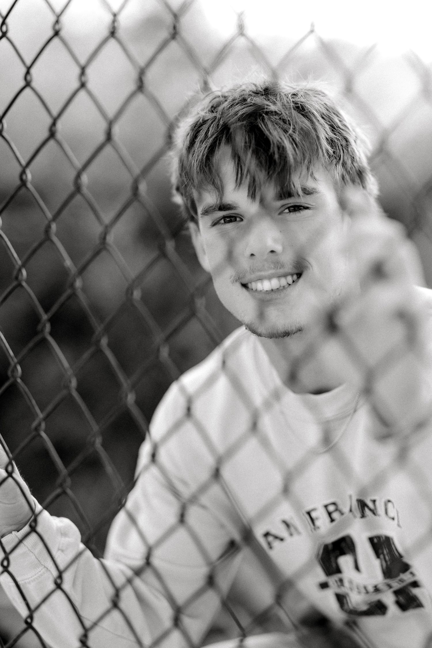Smiling young man leaning against a chain-link fence in a black and white portrait photo.
