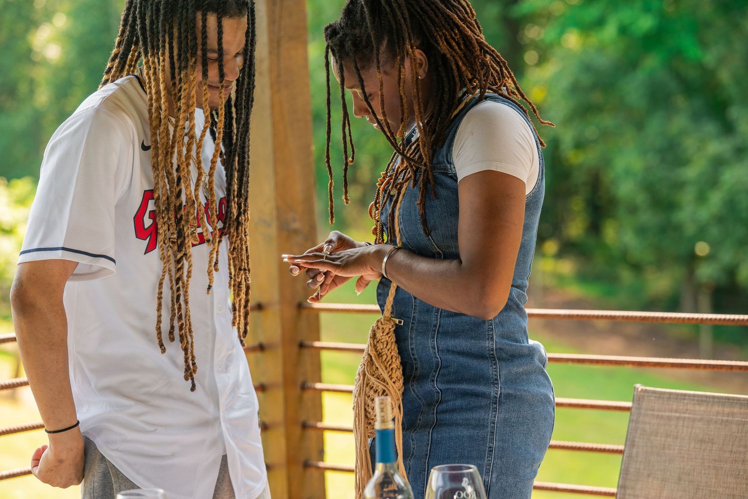 A couple with matching dreadlocks poses closely together on a wooden deck surrounded by greenery.