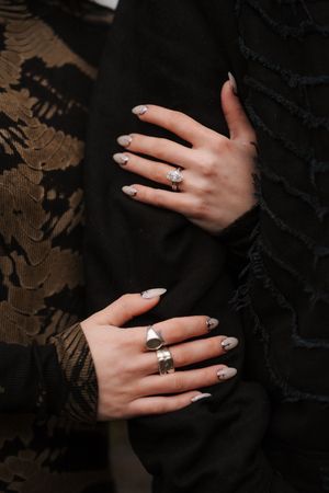Close-up of two hands with nude stiletto nails and silver rings, resting on black lace and fringe fabric.