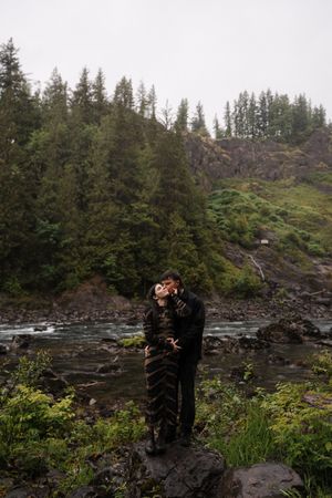 Couple kissing on rocks by a river surrounded by lush green forest and misty mountains in a romantic outdoor setting.