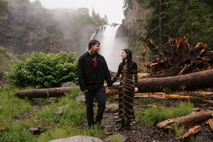 Couple holding hands near Snoqualmie Falls waterfall, surrounded by lush greenery and fallen logs on a misty day.
