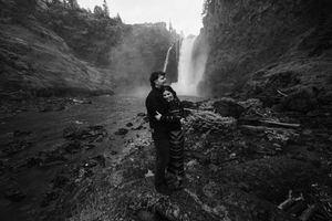 Couple embracing romantically in black and white photo before a stunning waterfall in a rocky canyon.