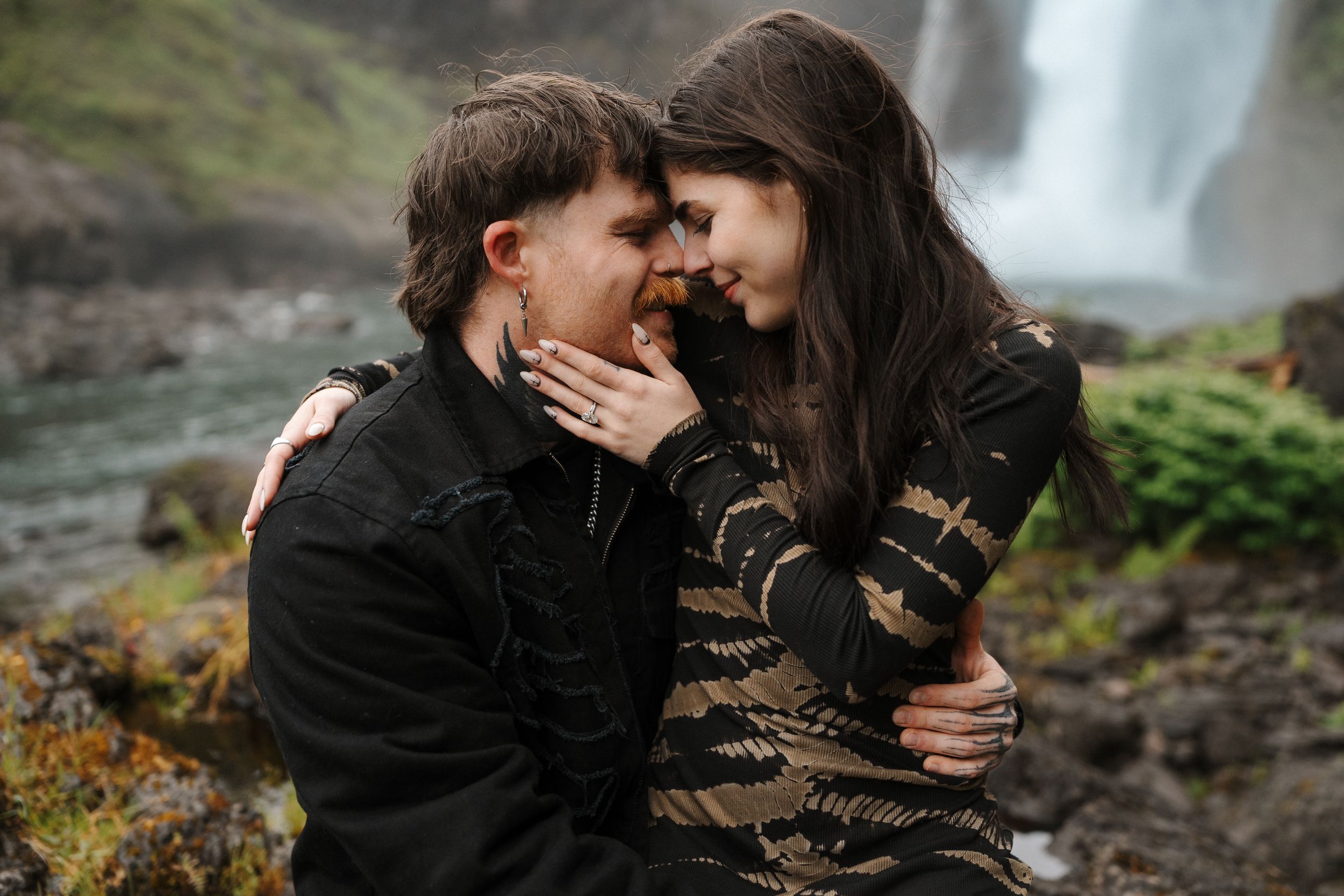 A couple shares a romantic moment in front of a waterfall, touching foreheads tenderly in nature.