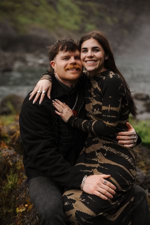 A smiling couple embraces outdoors near a misty waterfall, woman in patterned dress, man in black jacket.
