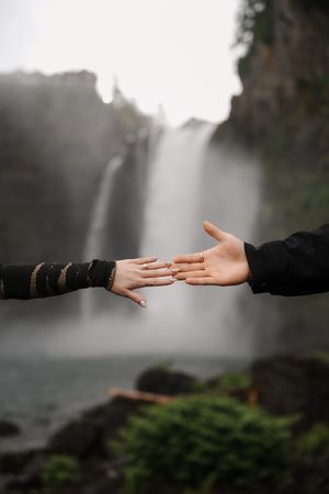 Two hands reaching toward each other in front of a misty waterfall, with one hand wearing a ring.