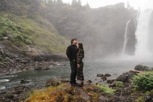 Couple sharing a romantic moment in front of a misty waterfall surrounded by lush greenery and rocky terrain.