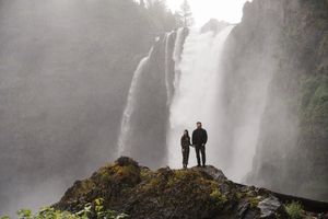 Two hikers stand on a rocky outcrop before a massive misty waterfall surrounded by lush green forest.