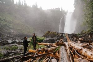 Two people stand on driftwood logs near a misty waterfall surrounded by lush green forest.