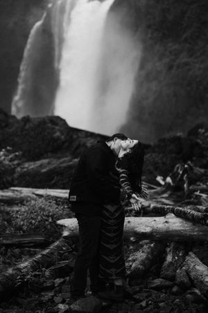 Black and white photo of a couple kissing in front of a dramatic waterfall surrounded by logs and lush greenery.